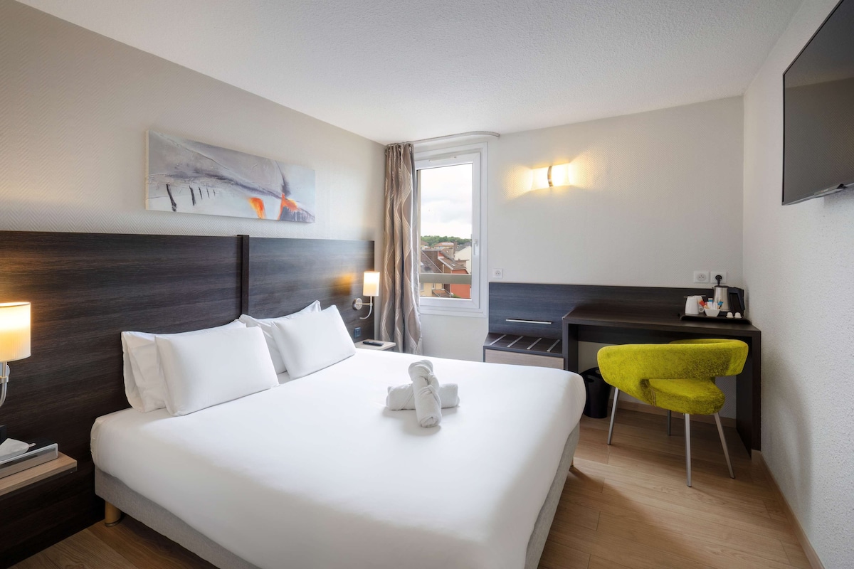 Una habitación de hotel con una cama grande, un escritorio, una silla amarilla brillante, un cuadro en la pared y un televisor montado en la pared.