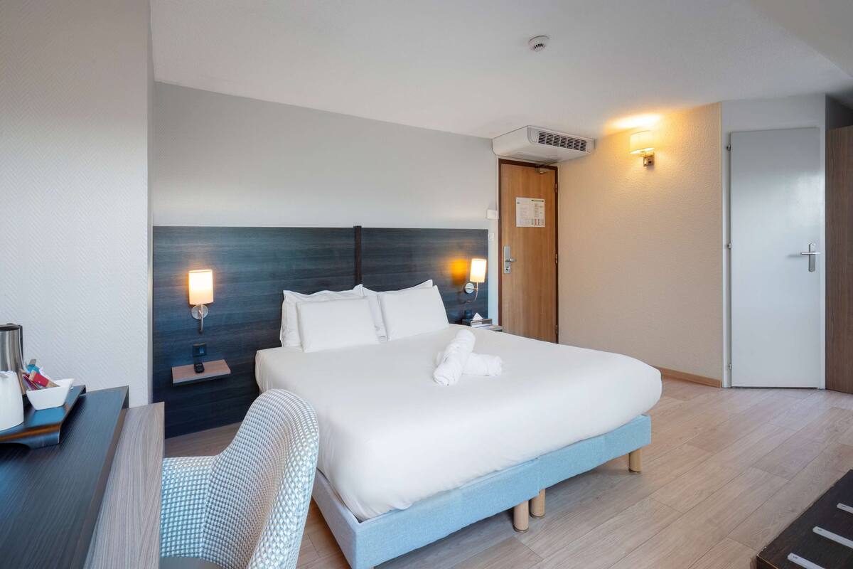 A modern hotel room with a large bed, bedside lamps, a desk with a chair, and a wooden door.