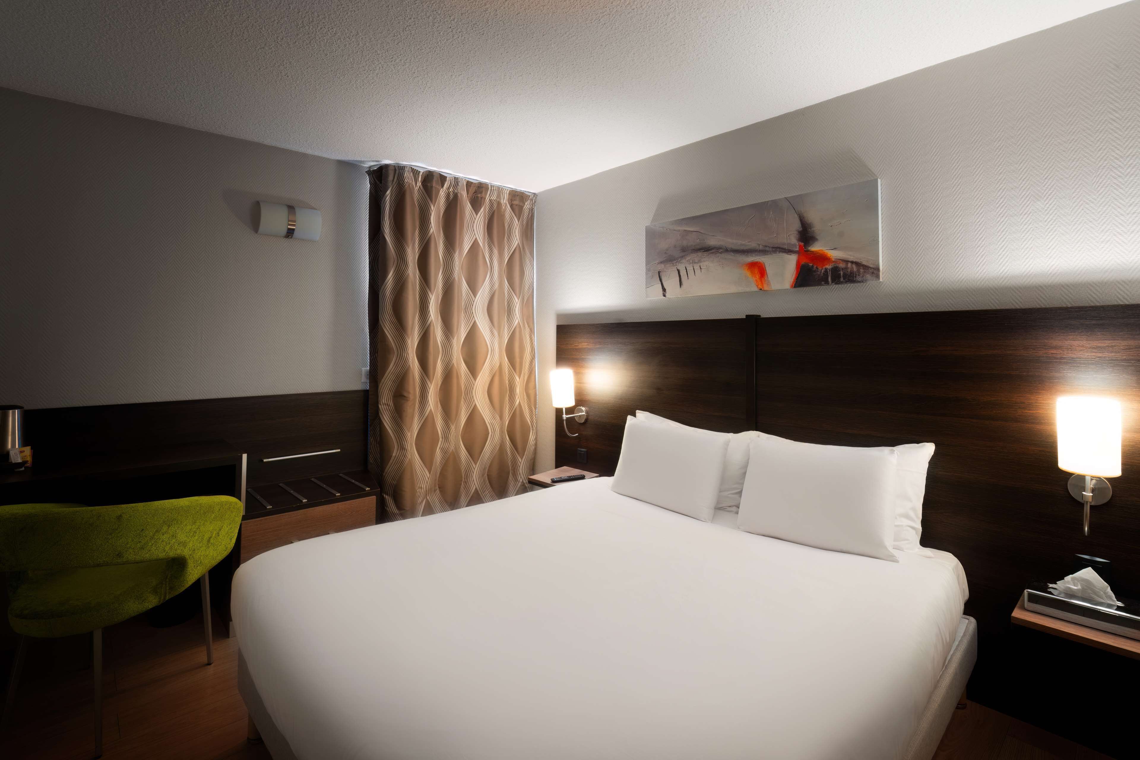 A hotel room with a large bed, a desk, a green chair, a nightstand with a lamp, and a painting on the wall.
