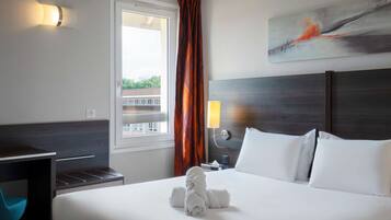 Quarto de hotel com cama bem arrumada, uma escrivaninha e uma cadeira. Há uma janela com cortinas, um quadro na parede e uma mesa de cabeceira com abajur.