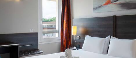 Habitación de hotel con una cama bien hecha, un escritorio y una silla. Se ve una ventana con cortinas, un cuadro en la pared y una mesita de noche con una lámpara.