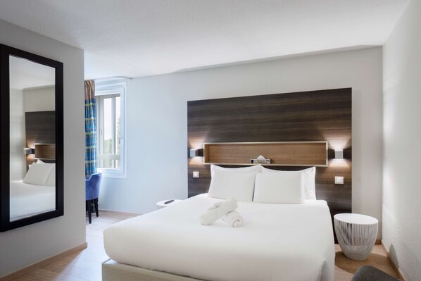 A modern hotel room with a large bed, a headboard with integrated lighting, a bedside table, a round side table, and a mirror.