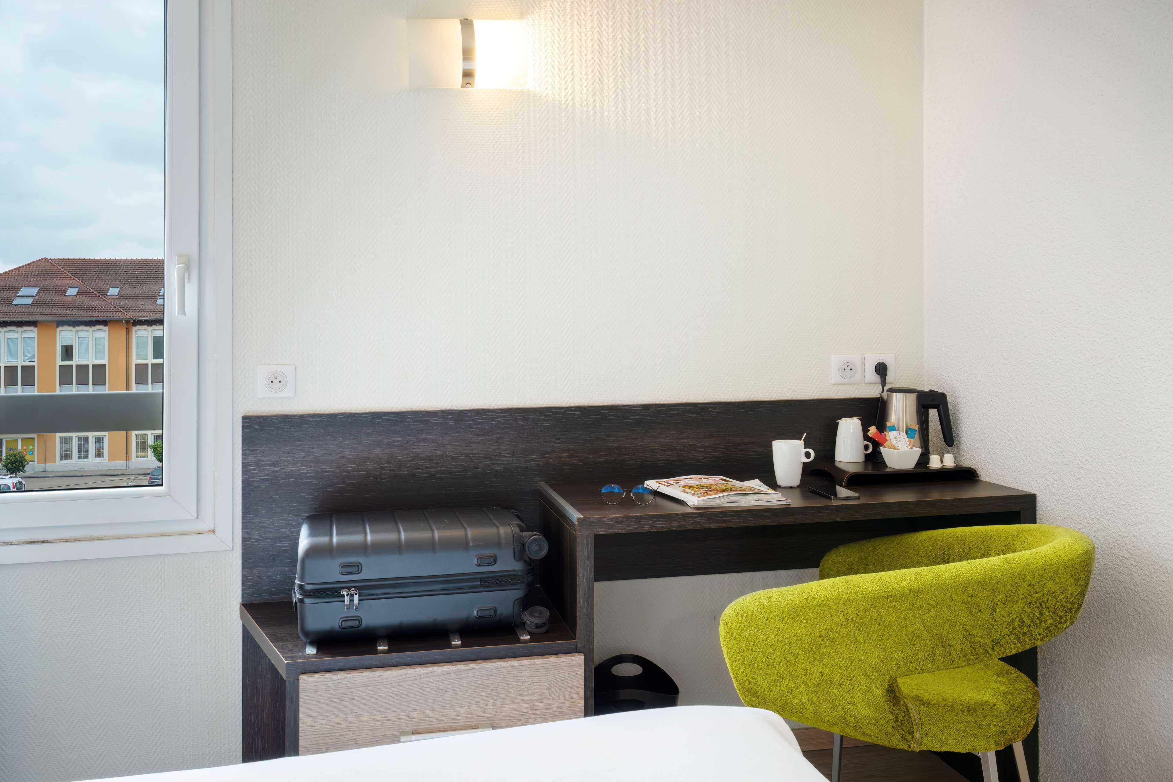 A hotel room with a bed, a desk with a suitcase, a bright green chair, and a window with a view of buildings.