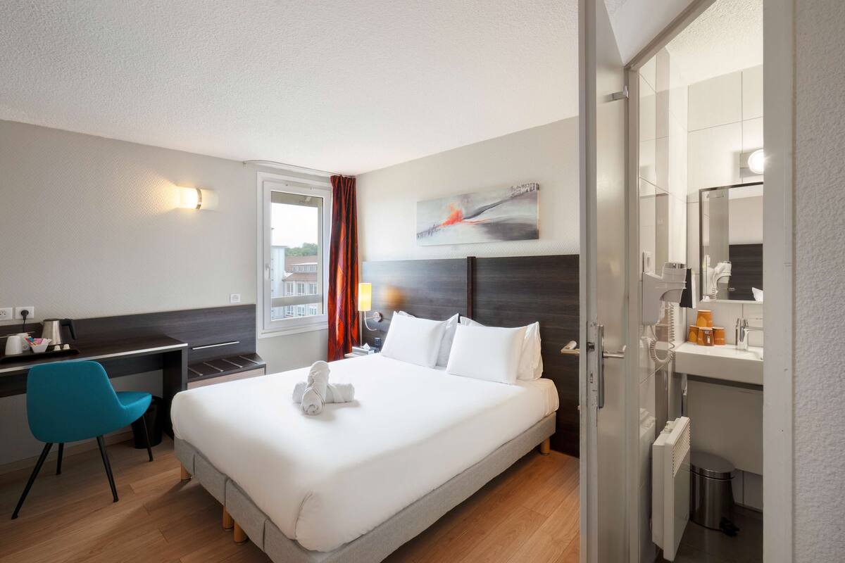 A hotel room with a bed, a desk, a chair, a window with curtains, a painting on the wall, and a bathroom with a mirror and sink.