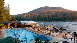 A pool area with lounge chairs and umbrellas, a dock with boats, and a forested hill in the background. - The Pines Resort (Bass Lake)