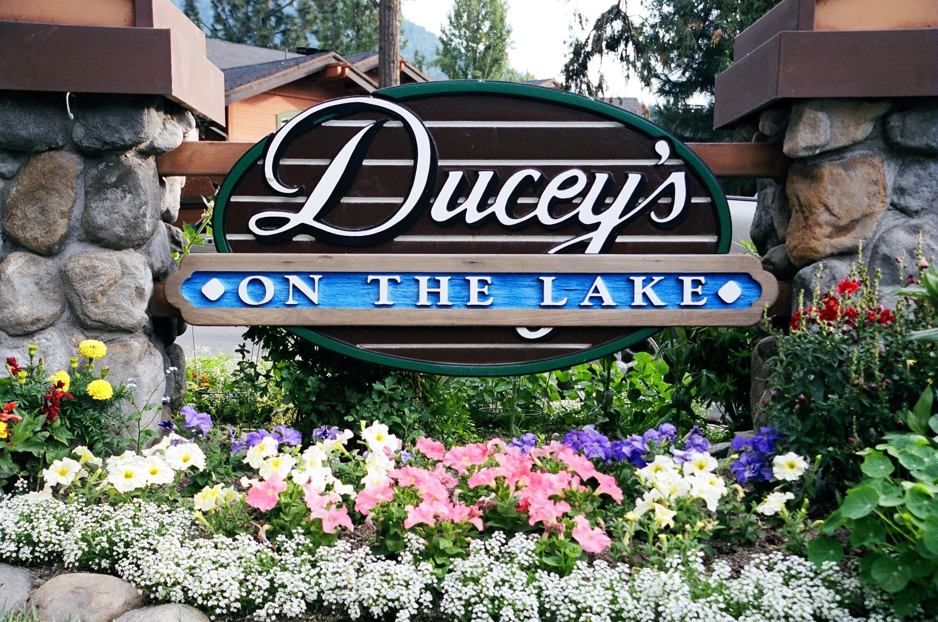 A sign for 'Ducey's On The Lake' with a stone wall and colorful flowers in front.