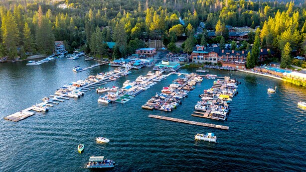 An aerial view of a marina with multiple docks and boats, surrounded by dense forest and mountains. - The Pines Resort (Bass Lake)