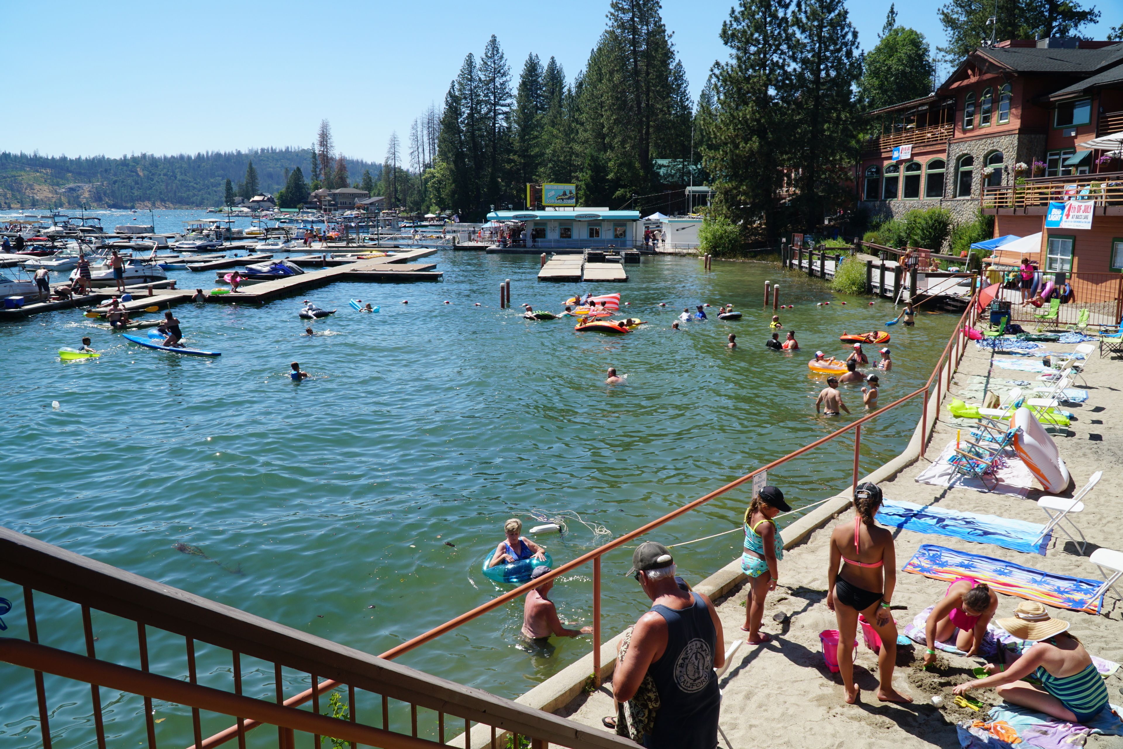 A lakeside scene with boats, people swimming, and a dock area with chairs and umbrellas.