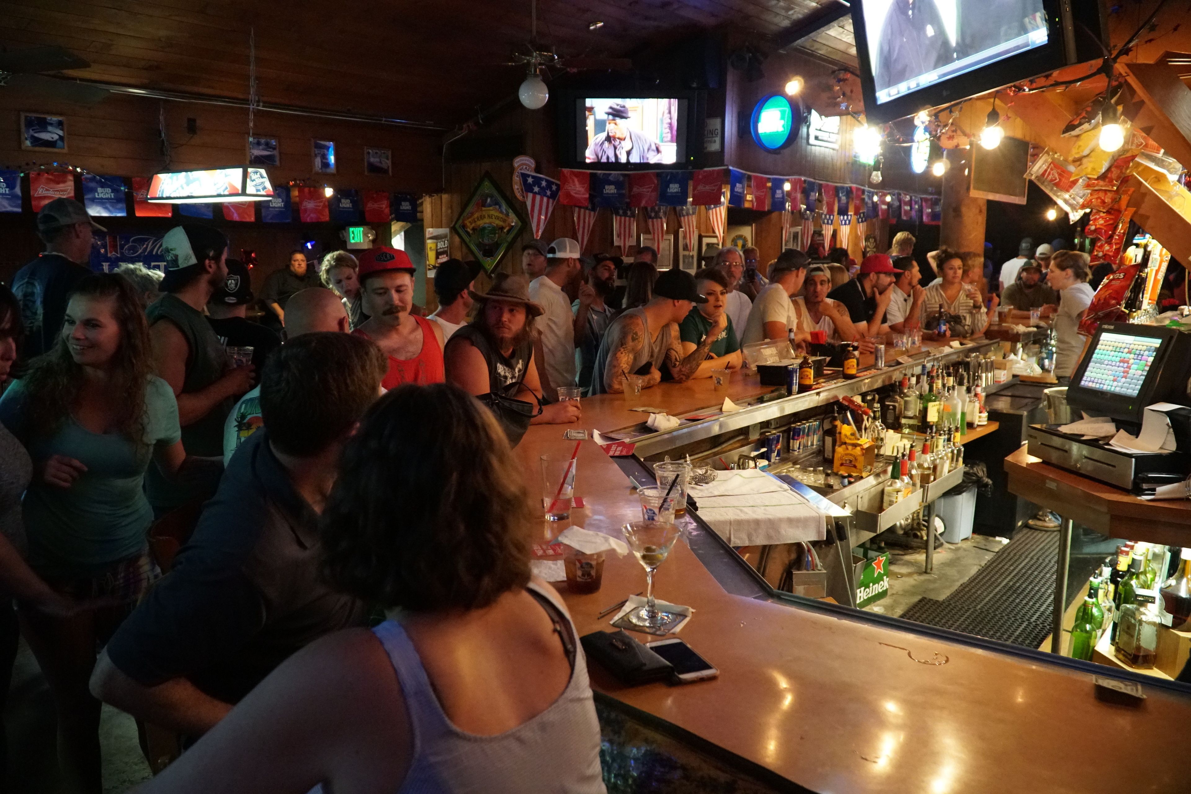 A crowded bar with a long counter, multiple televisions, and various flags.