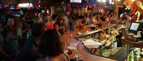 A crowded bar with a long counter, multiple televisions, and various flags.