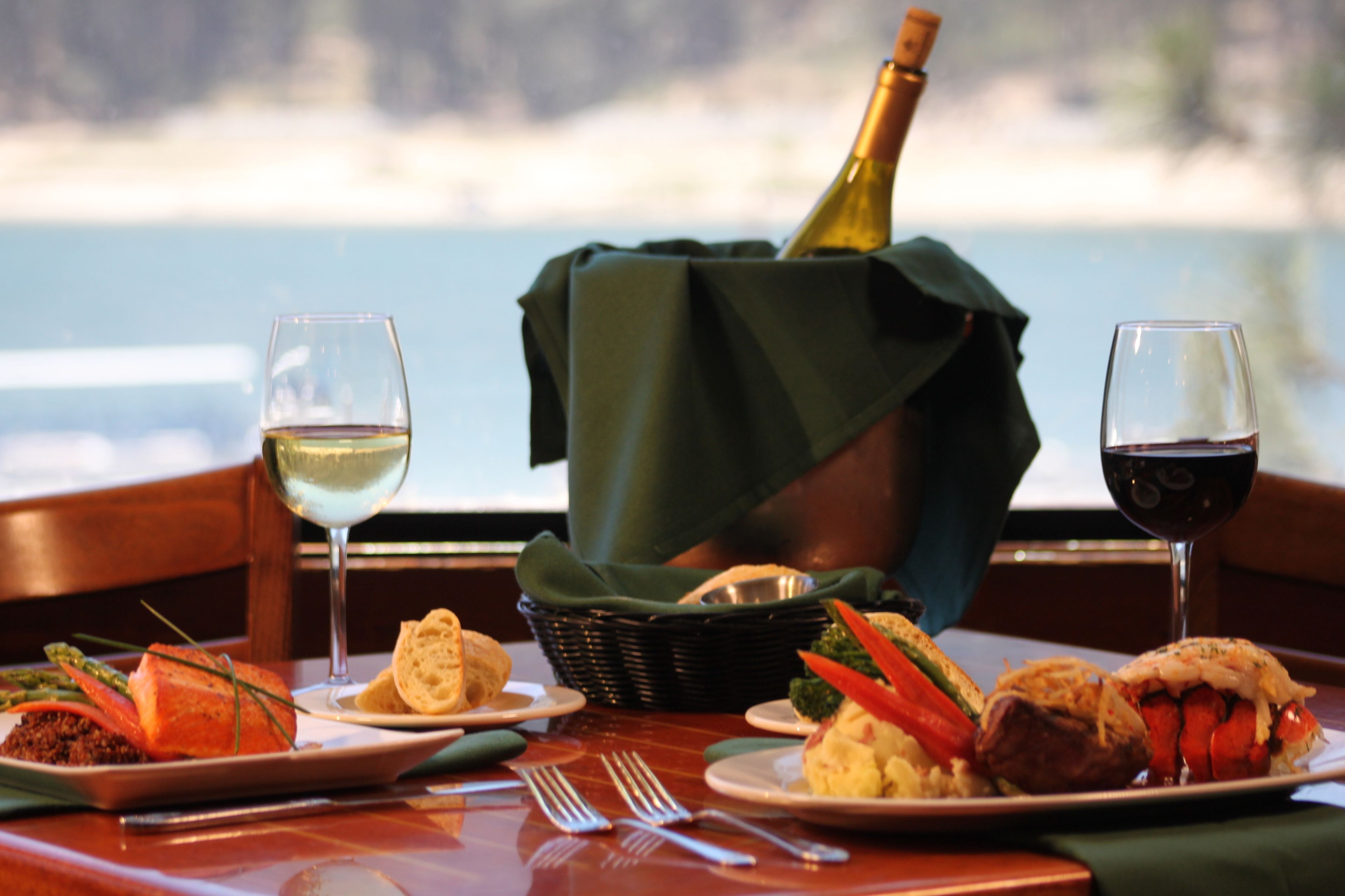 Una mesa dispuesta con platos de comida, dos copas de vino y una botella de champán, con vista al mar de fondo.