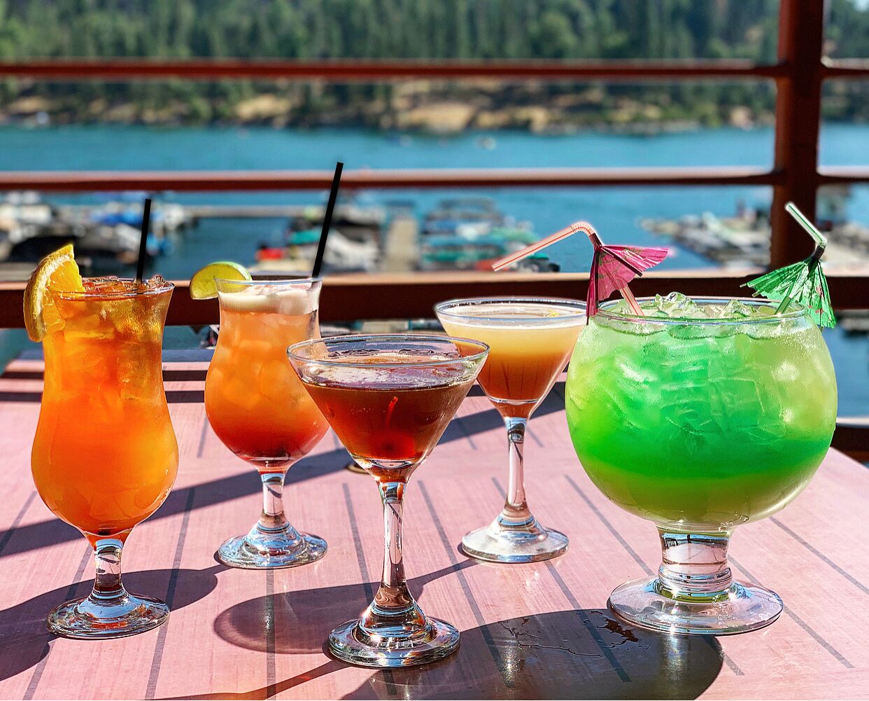 Ett bord med fyra färgglada cocktails, ett träbord och en utsikt över en småbåtshamn med båtar.