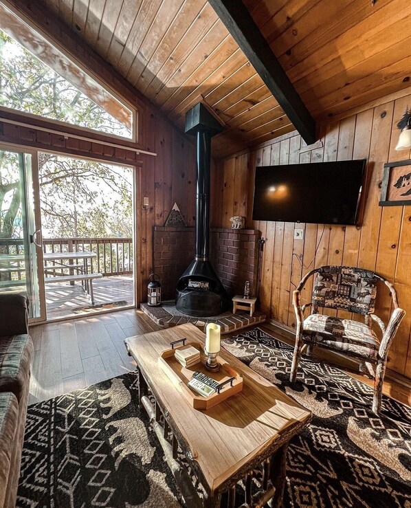 A cozy cabin interior with a wooden interior, a fireplace, a television, and a large window overlooking a deck.