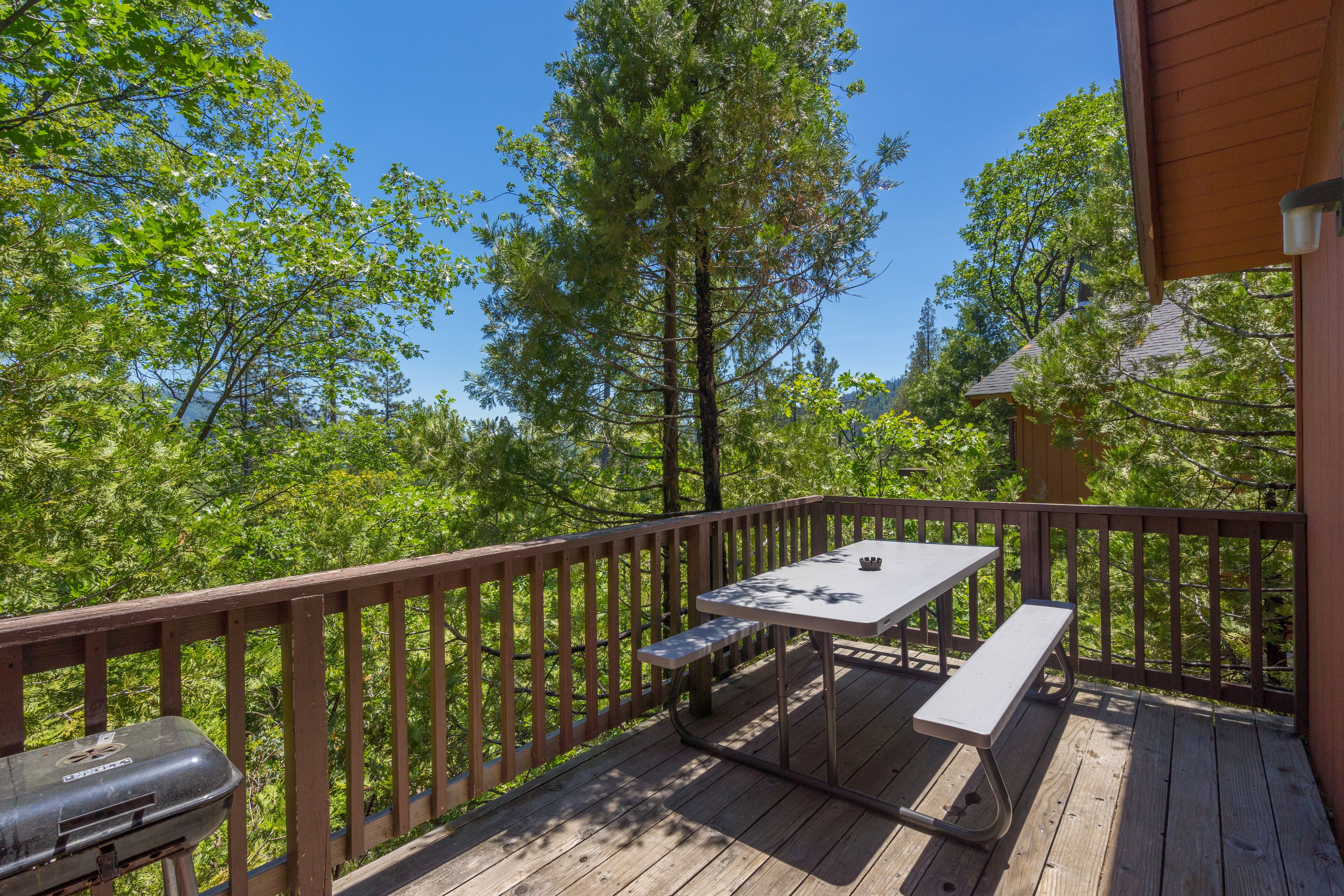 A wooden deck with a picnic table and bench, surrounded by trees and a clear sky.
