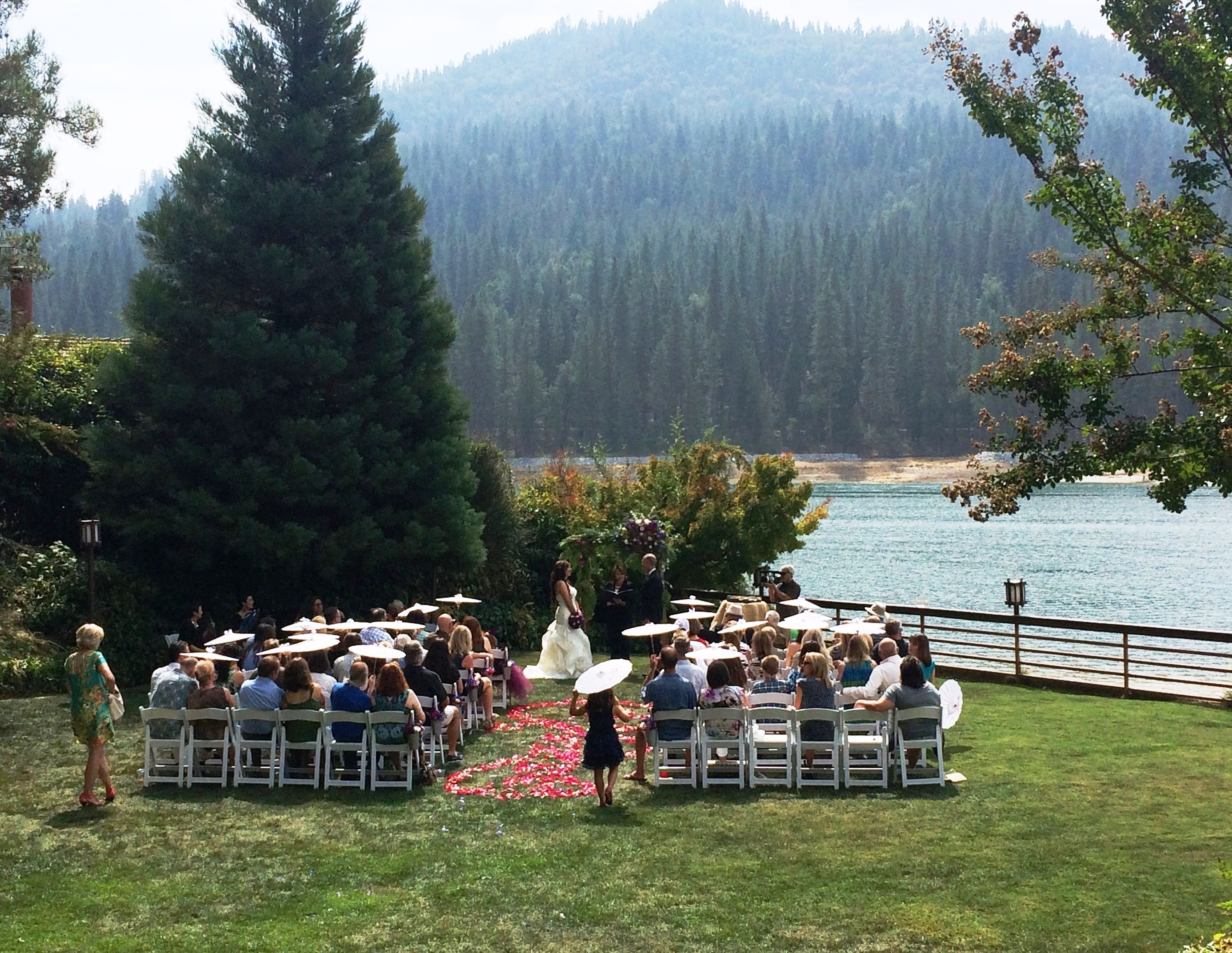 An outdoor wedding ceremony with guests seated on white chairs facing a couple walking down an aisle.