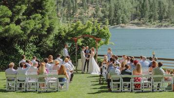 A wedding ceremony outdoors with guests seated on white chairs facing a wooden arch.
