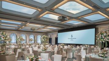 A spacious banquet hall with tiered seating, large windows, and a central screen displaying the hotel name.