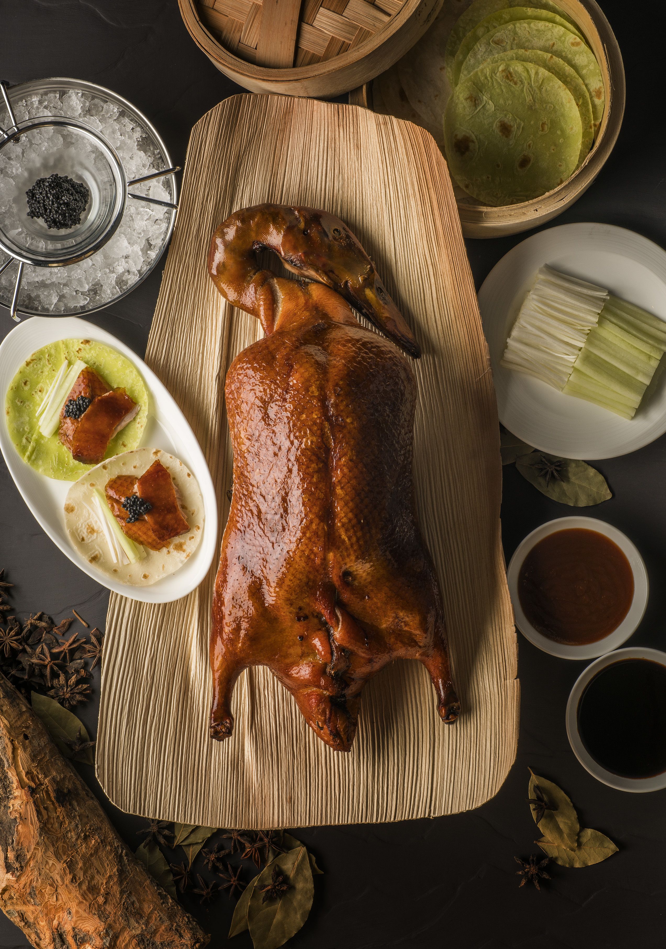 Un pato asado sobre tabla de madera, con un tazón de salsa, un plato de tortillas y un tazón de caviar.