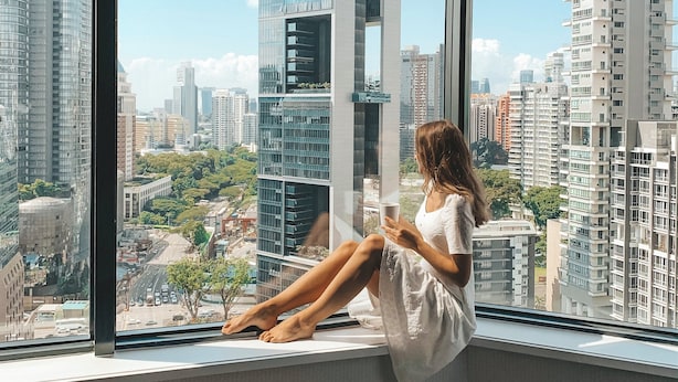 A woman sitting by a large window with a city view, a vase with flowers, and a small table. - Four Seasons Hotel Singapore (Singapore)