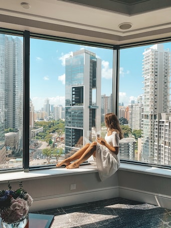 A woman sitting by a large window with a city view, a vase with flowers, and a small table.