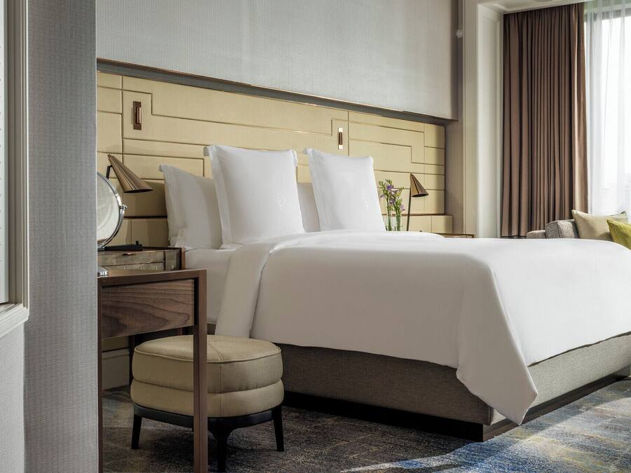 Et hotelværelse med seng, natbord, lampe, stol og udsigt over byen fra vinduet.