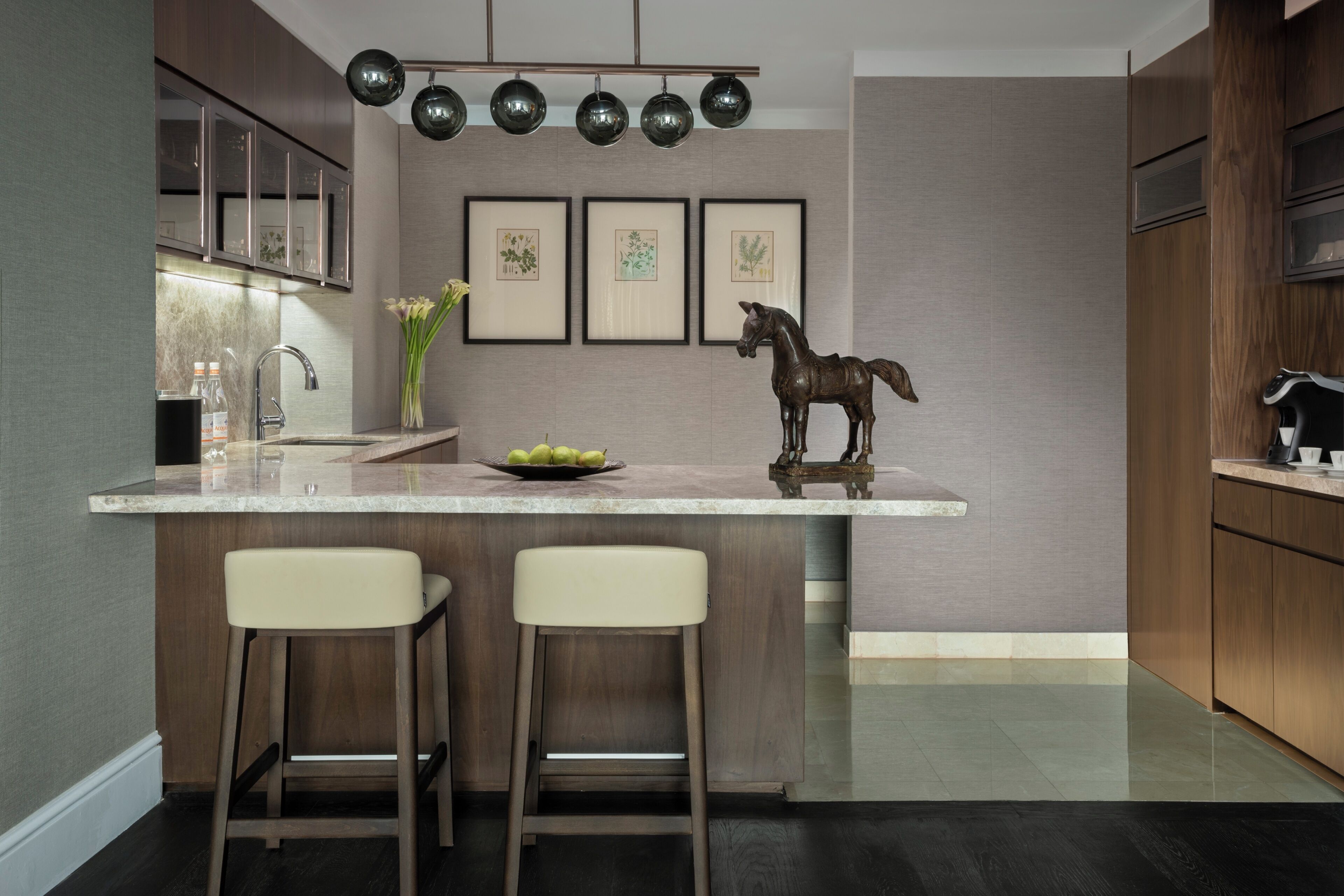A modern kitchen with a marble countertop, two bar stools, a decorative horse sculpture, and framed botanical prints on the wall.