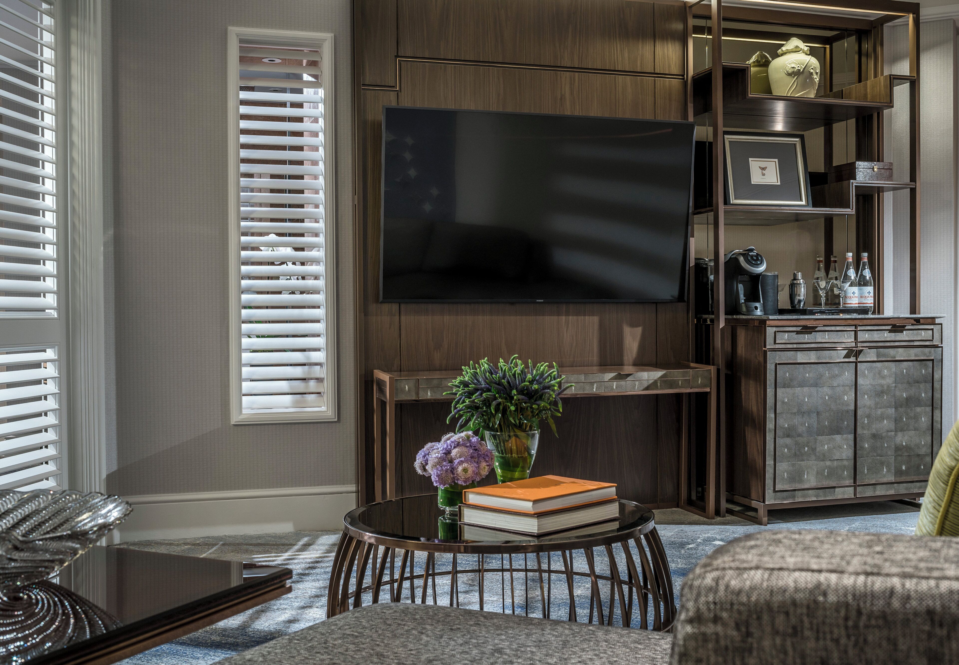 Uma sala de estar moderna com uma televisão de tela plana na parede, uma mesa de centro com livros e um vaso de flores, e uma área de estar com um sofá.