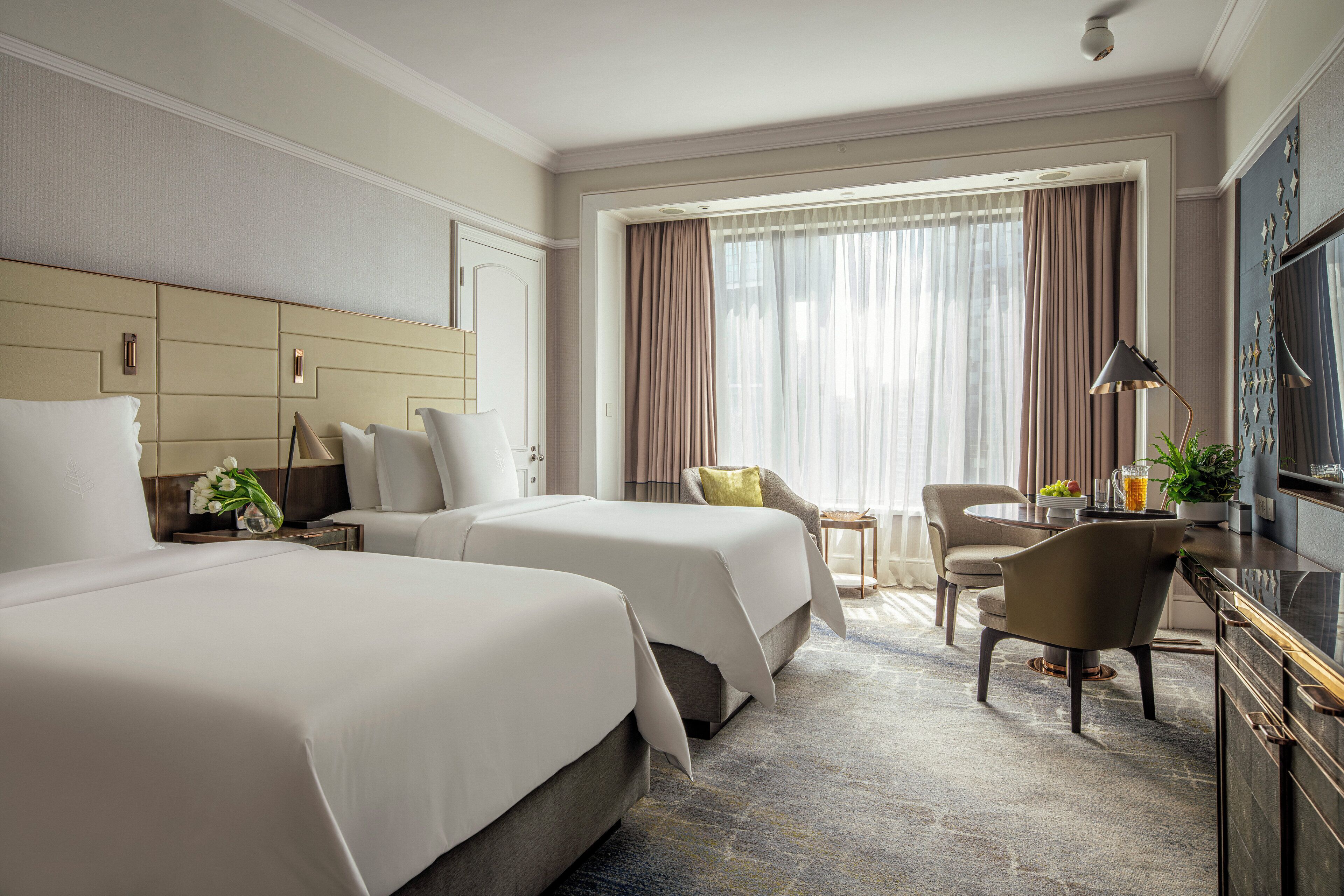 A hotel room with two beds, a desk, a chair, a TV, and a large window with curtains.