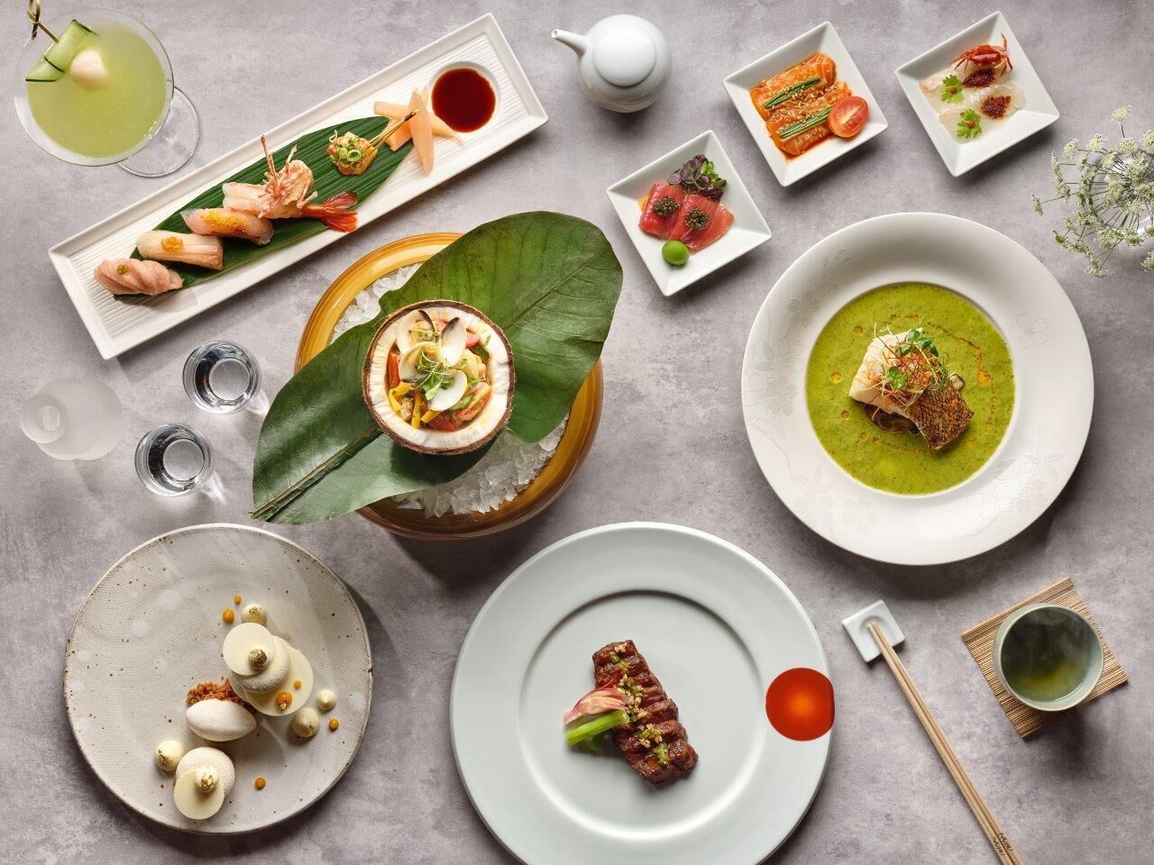 A variety of dishes including sushi, sashimi, and a green soup, arranged on a table with a leaf.