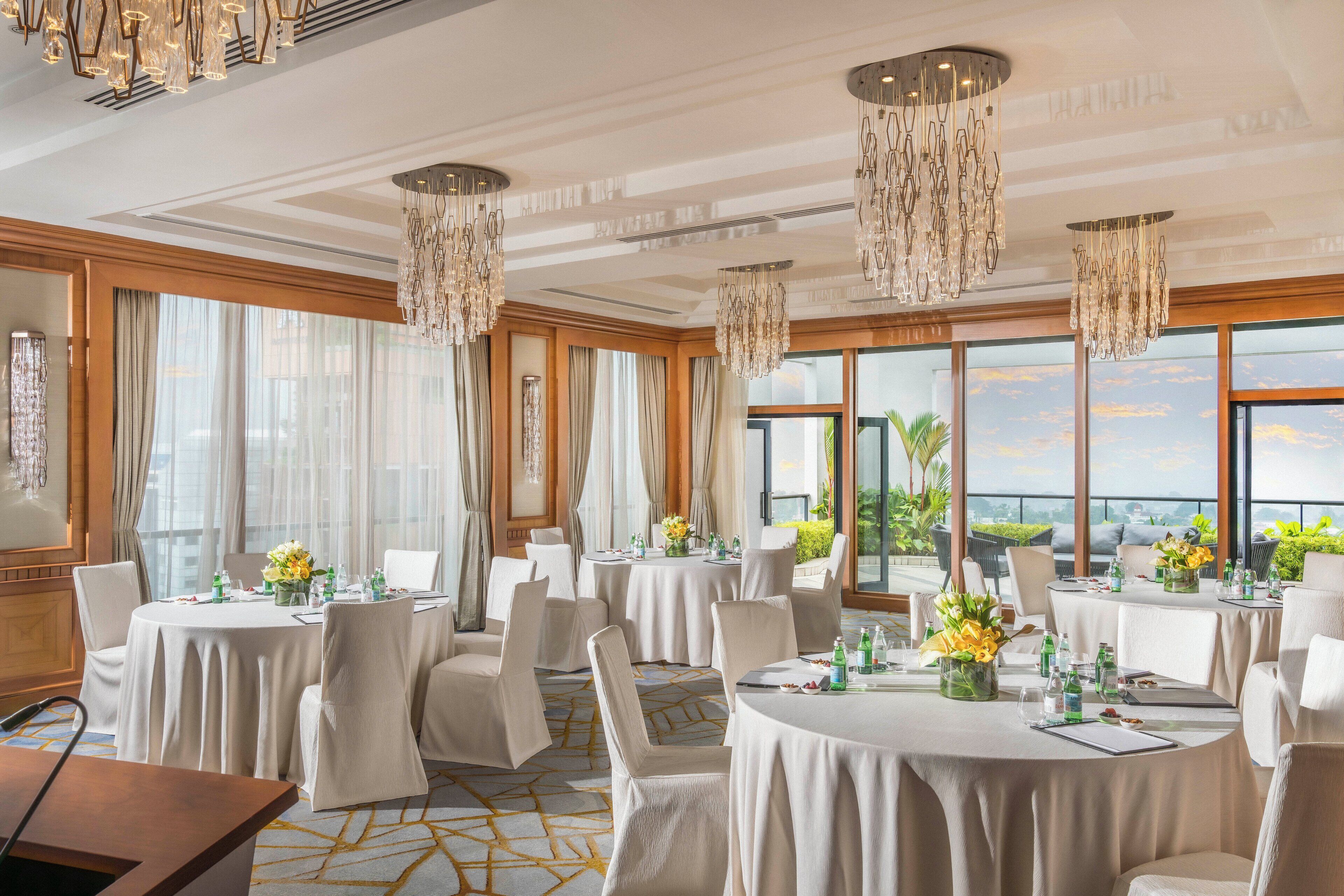 A hotel dining area with round tables, white chairs, chandeliers, and large windows with a view.
