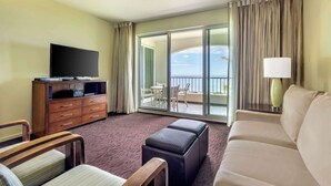 Une chambre d’hôtel équipée d’un téléviseur à écran plat posé sur un meuble en bois, d’un canapé beige, d’un fauteuil en bois, d’un petit pouf, d’un ventilateur de plafond et d’une grande porte-fenêtre coulissante donnant sur un balcon avec vue sur l’océan.