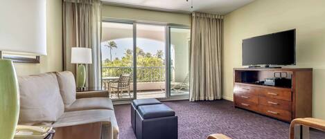 A hotel room with a sofa, ottoman, TV, and a balcony with curtains.