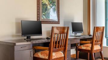 A workspace with two desks, each with a computer monitor, keyboard, and mouse. There are two wooden chairs with patterned cushions. A framed tropical artwork is on the wall above the desks.