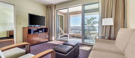 A hotel room with a large flat-screen TV, a sofa, a loveseat, and a balcony with a view of palm trees.