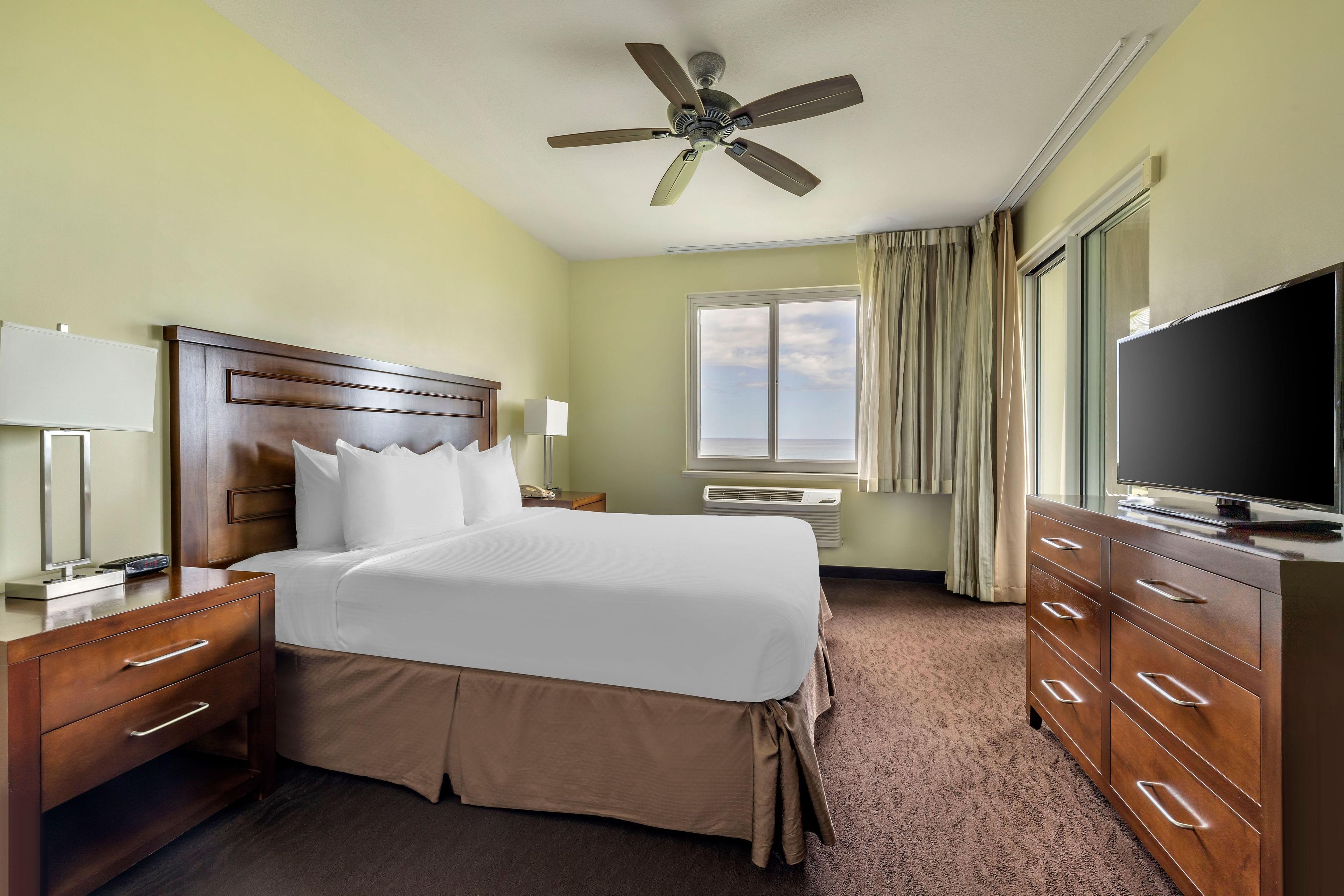 Ein Hotelzimmer mit Bett, Schreibtisch, Fernseher, Deckenventilator und einem Fenster mit Vorhängen.