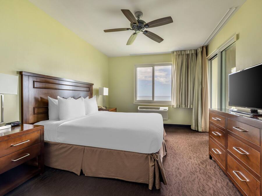 A hotel room with a bed, a desk, a television, a ceiling fan, and a window with curtains.