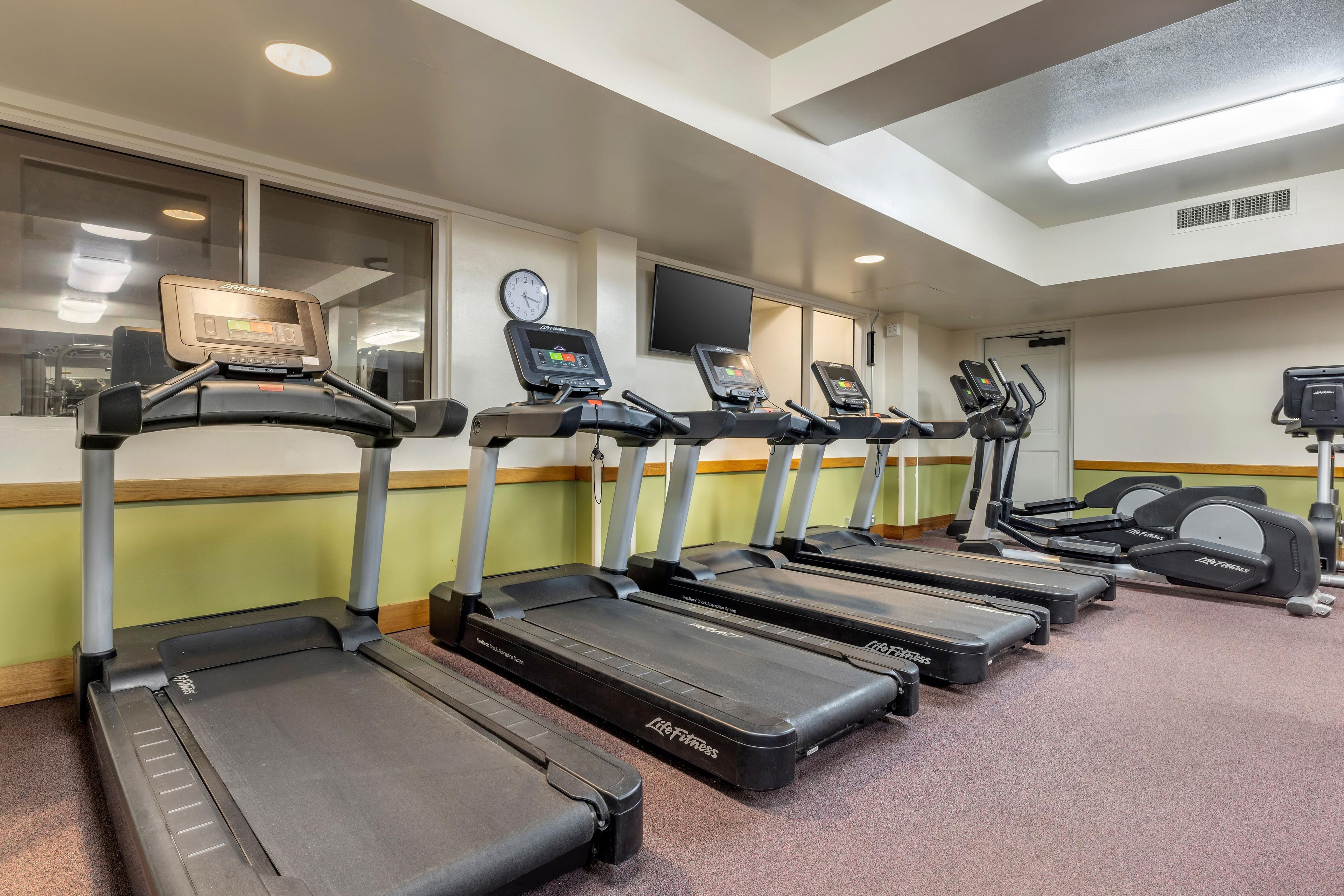 Centro de fitness com esteiras, relógio na parede e televisão.