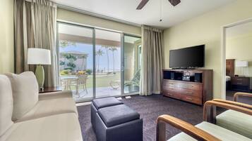 A hotel room with a sofa, ottoman, flat-screen TV, wooden cabinet, and a view of a pool and palm trees.