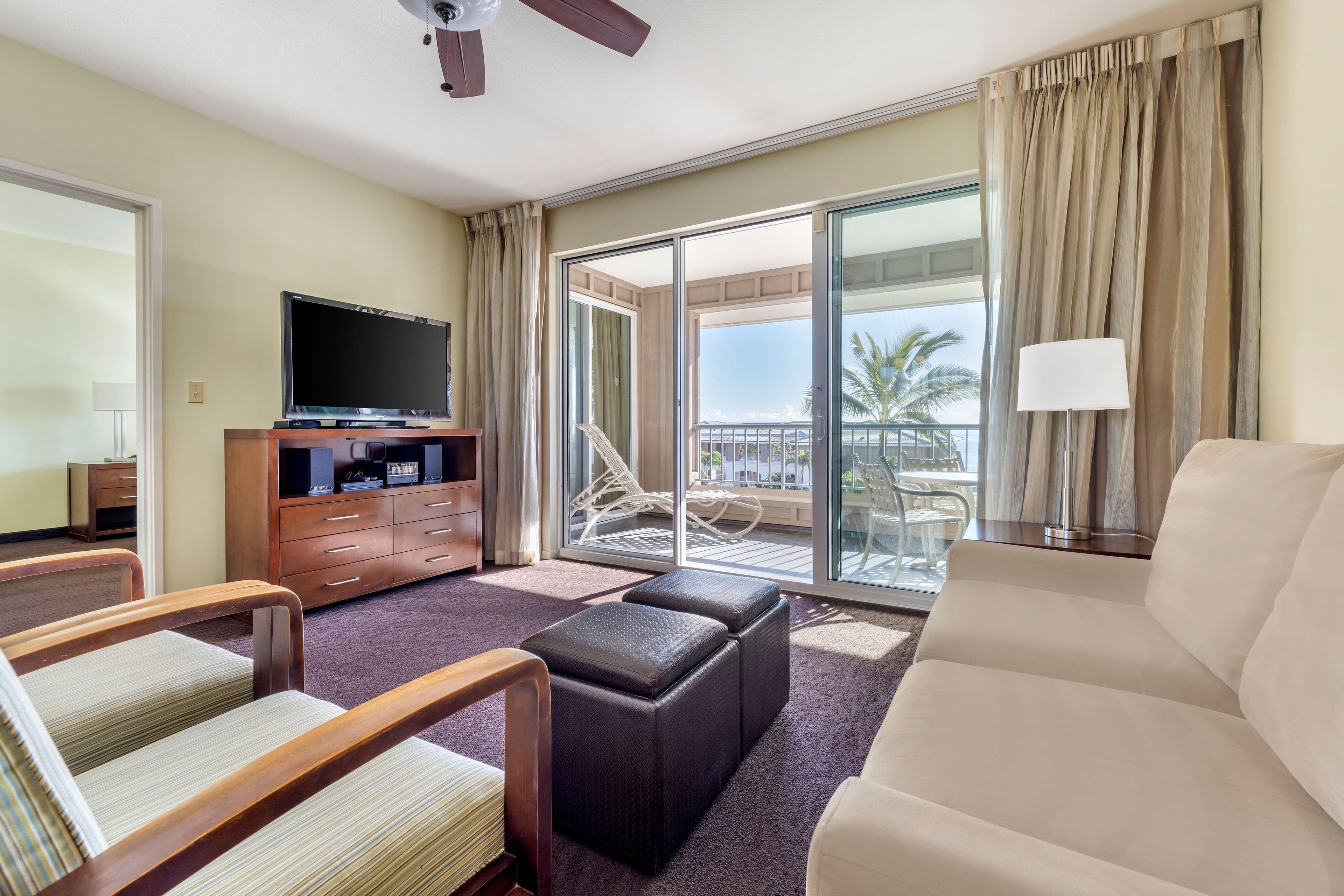A hotel room with a flat-screen TV, a sofa, a chair, a small table, a lamp, and a balcony with a view of palm trees.