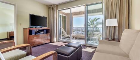 A hotel room with a flat-screen TV, a sofa, a chair, a small table, a lamp, and a balcony with a view of palm trees.