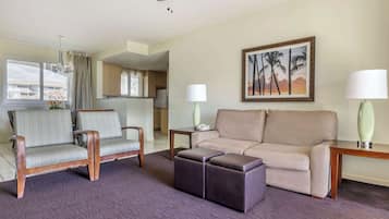A hotel room with a sofa, armchairs, a coffee table, a painting of palm trees, and a view of the kitchen.