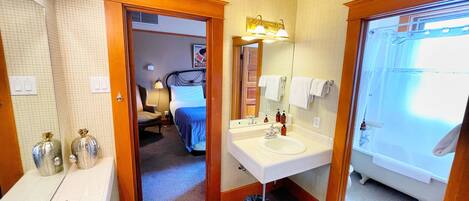 Un baño de hotel con un lavamanos grande, un espejo y una ducha. Se ve una cama en la habitación contigua.
