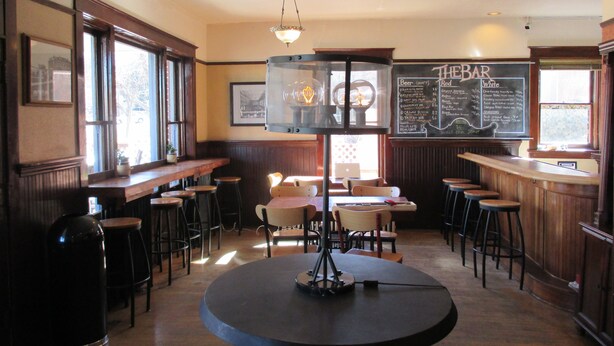 Eine gemütliche Bar mit Holztresen, Barhocker und einem runden Tisch mit einer Dreifußlampe. Eine Kreidetafel an der Wand zeigt eine Auswahl verschiedener Biersorten.