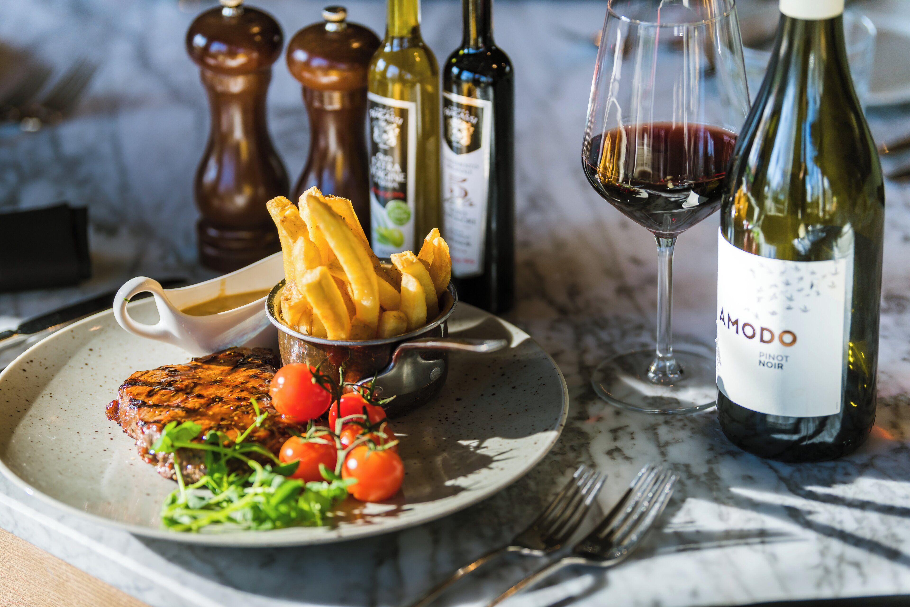 A plate with steak, fries, and vegetables, a glass of red wine, and two bottles of wine on a table.