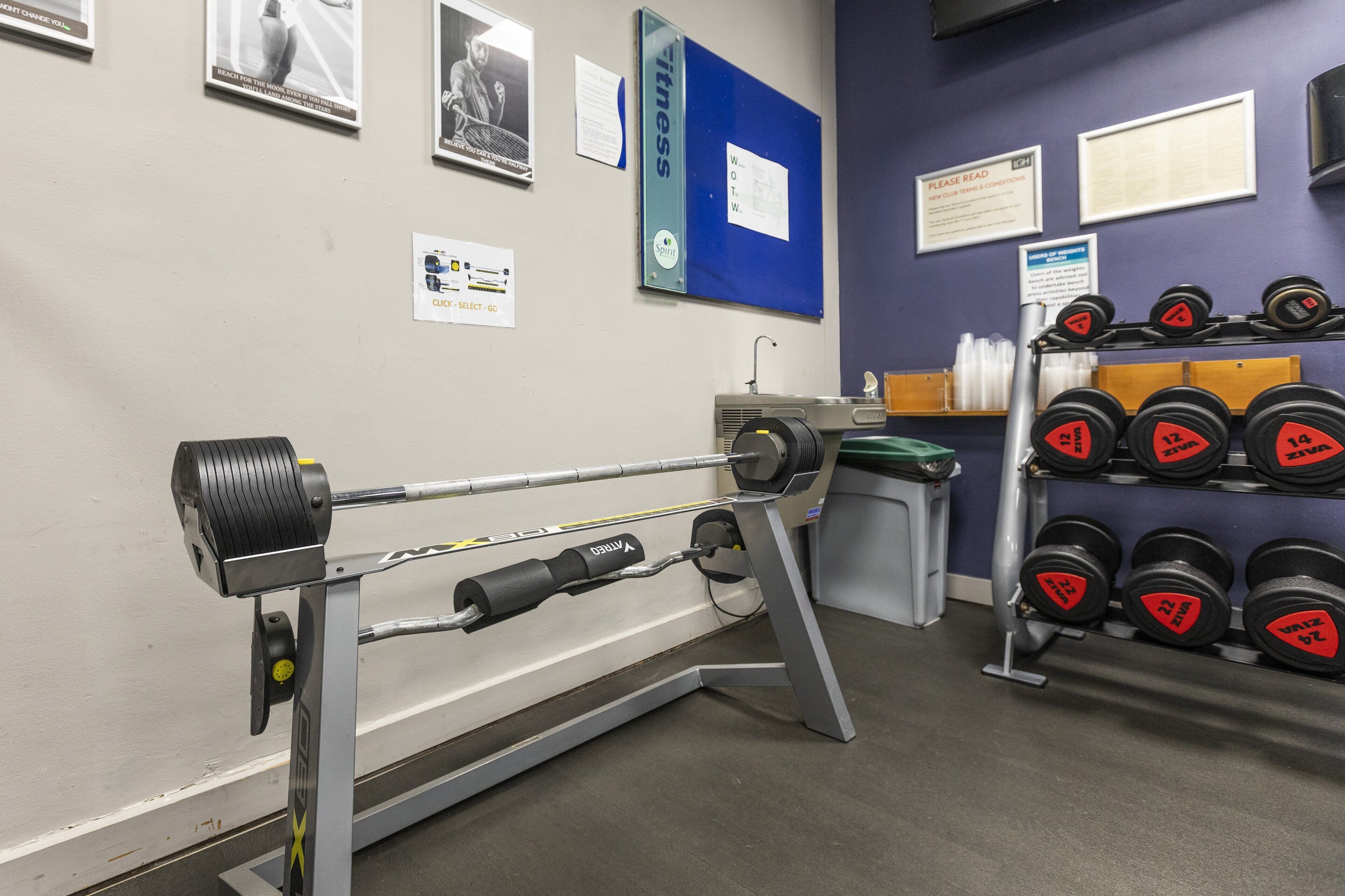 Ein Fitnessraum mit Krafttrainingsgeräten, darunter eine Langhantel und Kurzhanteln.