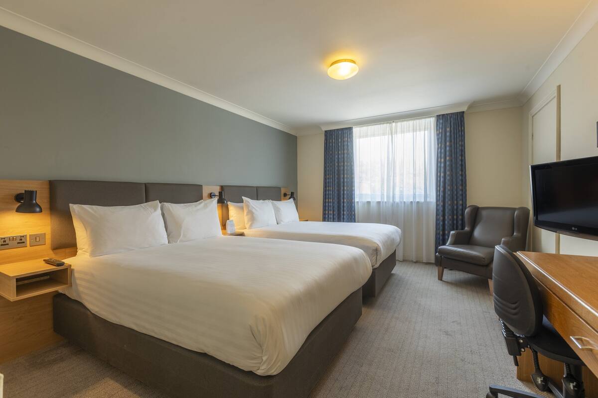 Une chambre d’hôtel avec deux lits, un bureau avec une chaise, une télévision et une fenêtre avec des rideaux.