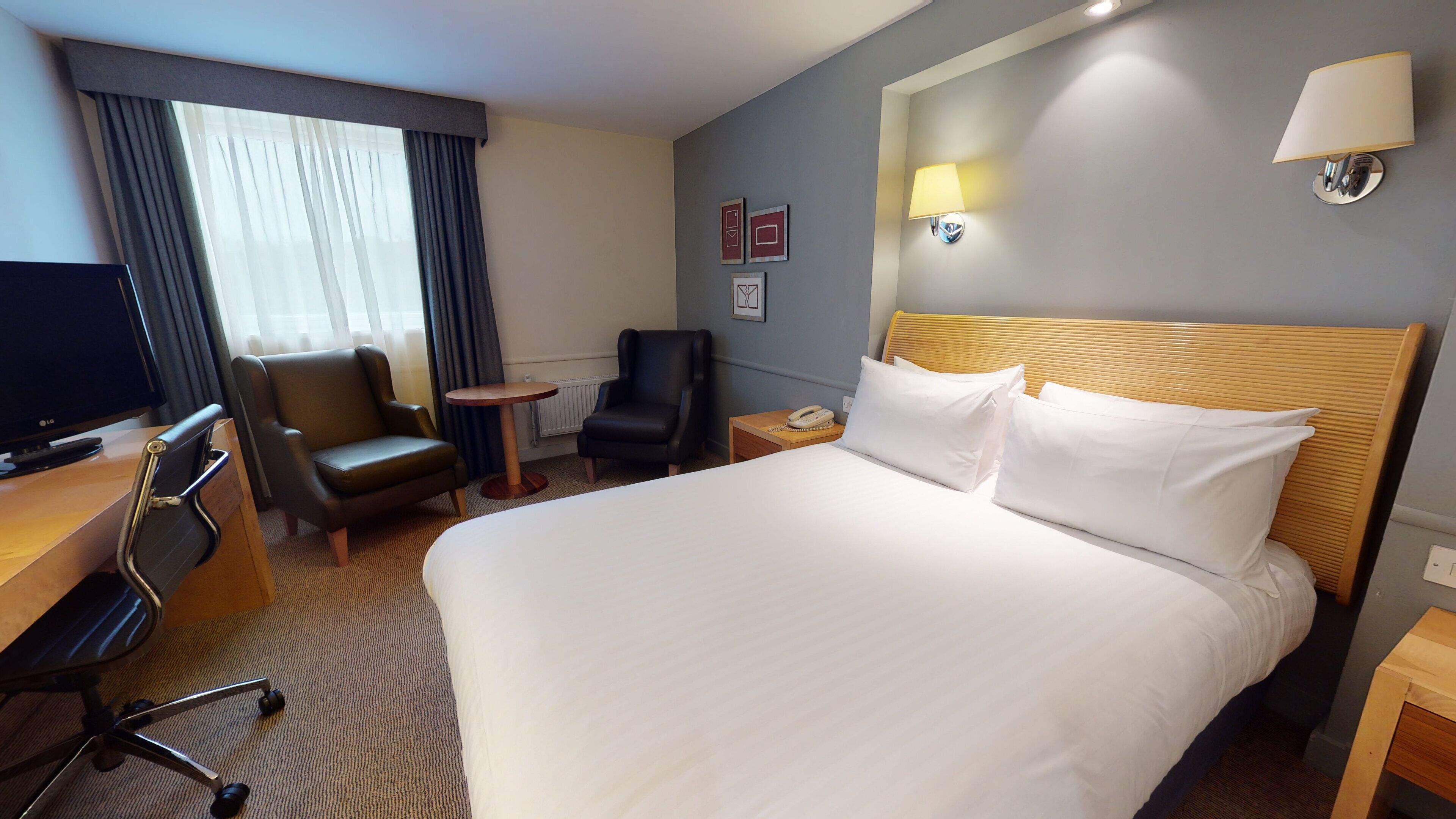 A hotel room with a large bed, a desk with a computer, a chair, a small table, and a lamp.