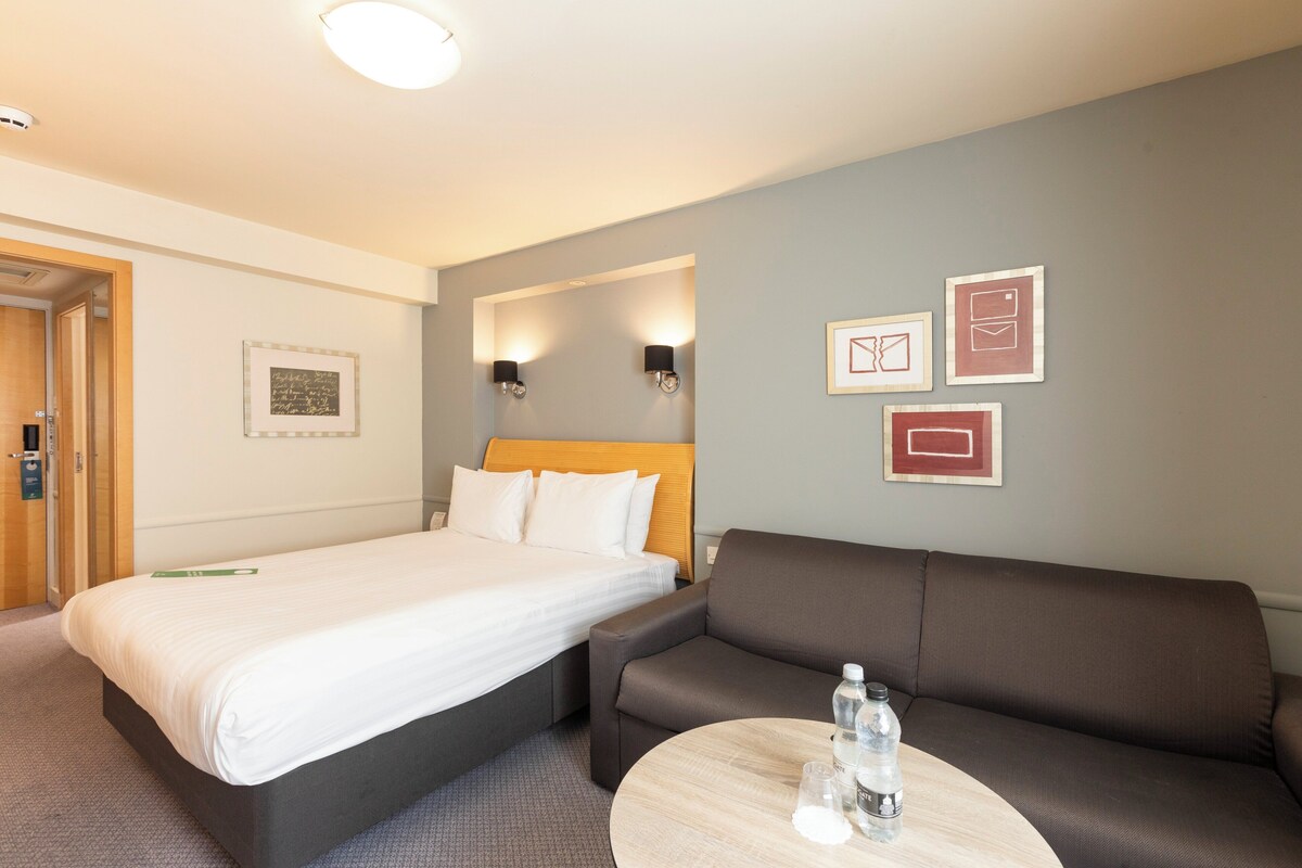 Une chambre d’hôtel avec un lit, un canapé, une table ronde et des lampes fixées au mur.