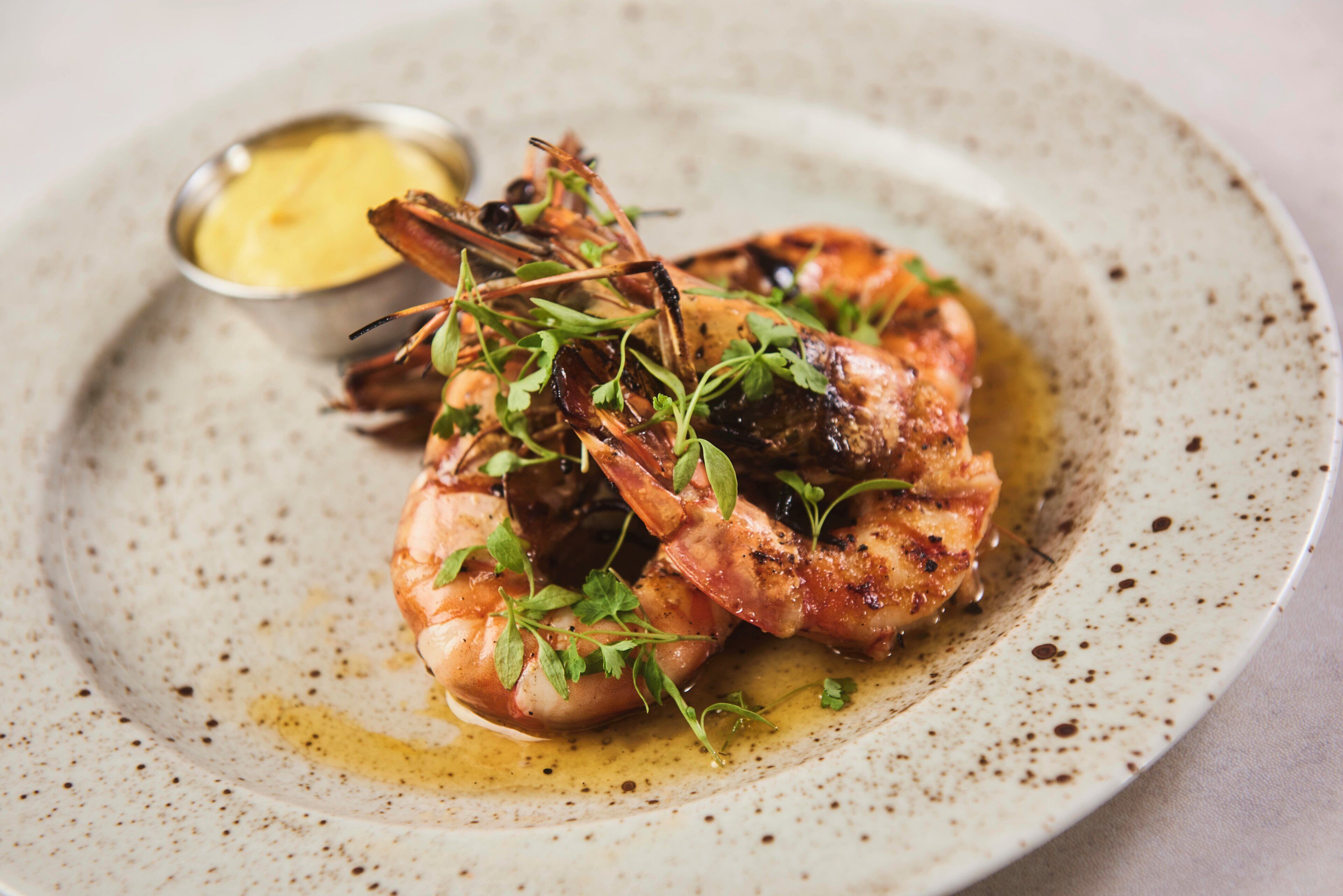 A plate with grilled shrimp and greens, accompanied by a small bowl of sauce.