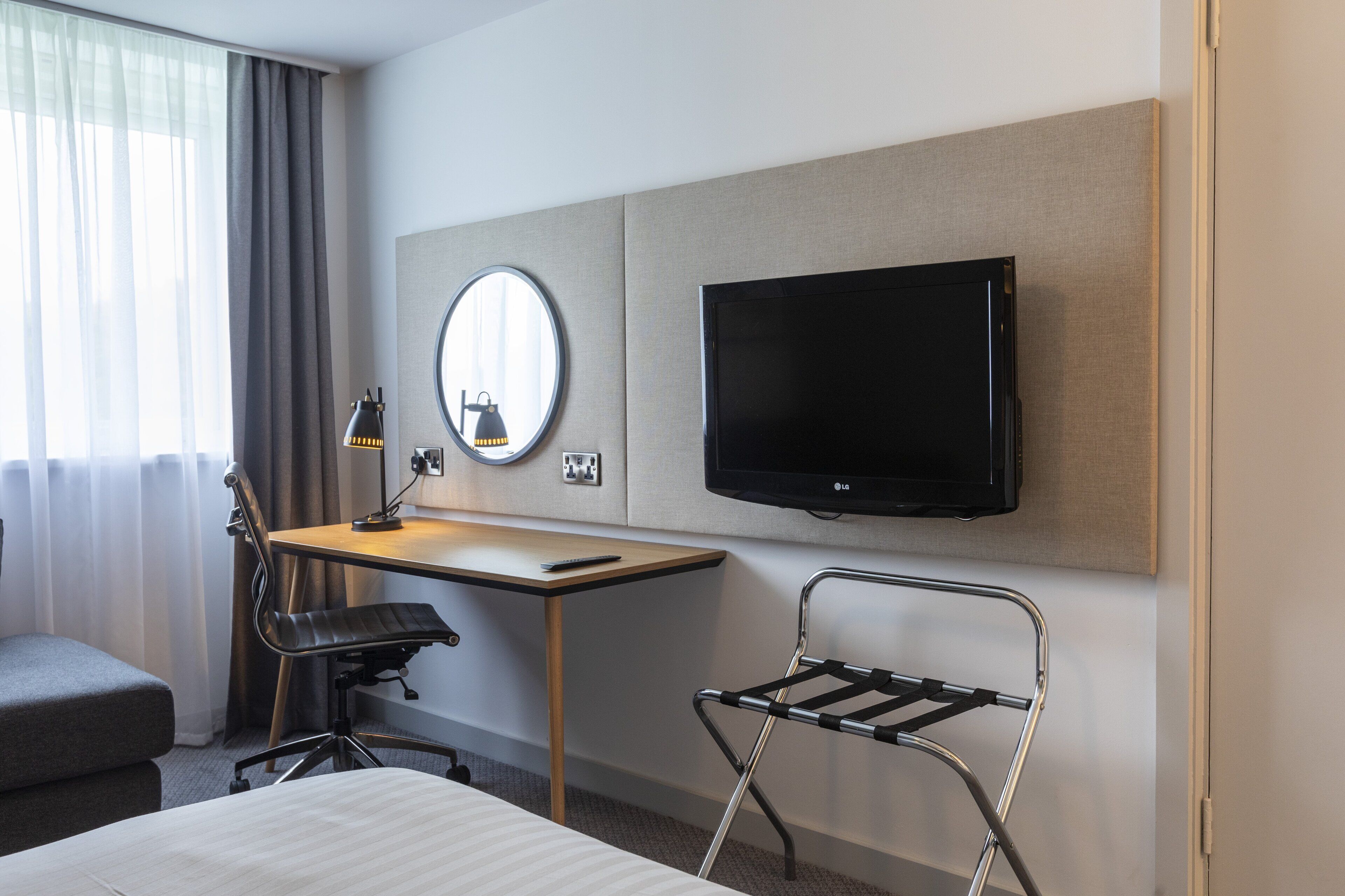 A hotel room with a bed, a desk with a chair, a TV mounted on the wall, a mirror, and a window with curtains.